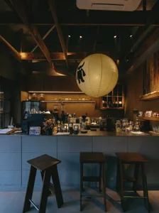 Interior view of a specialty coffee shop with dark wood stools, concrete counter, wooden ceiling beams, and a large illuminated Japanese paper lantern with a bull logo.