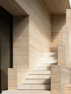 Close-up view of a modern monolithic staircase constructed from beige travertine stone with strong natural light creating sharp shadows on the textured wall and steps.