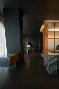Modern dark interior featuring a large green marble kitchen island, a tall brown onyx pillar, floating wood console, and a distant dining area with pendant light.