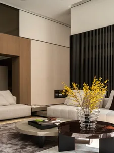 Contemporary luxury living room featuring a beige L-shaped sectional sofa, round coffee tables in dark wood and light stone, a dark vertical slatted accent wall, and a large glass vase with yellow flowering branches.