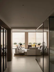 Modern minimalist living room featuring light travertine walls and floor, neutral fabric sofas, and large glass doors opening to a landscaped garden with trimmed shrubs.