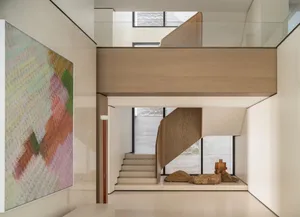 Modern minimalist double-height foyer with a curved oak staircase, large colorful abstract painting, stone sculptures, and high windows allowing natural light.