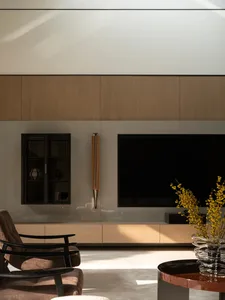 Modern minimalist living room featuring a large oak wood panel entertainment wall, wall-mounted TV, dark wood display cabinet, brown upholstered armchair with black frame, and a glossy round coffee table with yellow forsythia branches.