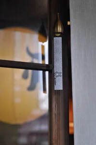 Close-up of a Japanese noren curtain with a black wave pattern hanging next to a brass wind bell on a dark wooden door frame, with a blurred paper lantern in the background.