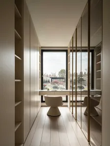 Modern narrow home office featuring built-in beige lacquer cabinetry, a large picture window overlooking a landscaped garden, a white bouclé swivel chair, and brass-framed glass doors.