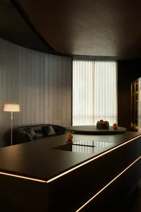 Modern dark interior featuring a curved wooden ceiling, vertical grey wall panels, black leather sofa, round dining table, and a long kitchen island with integrated LED lighting and induction cooktop.