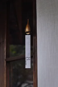 A brass teardrop-shaped wind chime with a white patterned paper strip hanging in front of a dark wooden Japanese-style window with blurred greenery outside.