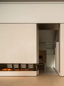 Modern minimalist interior featuring a long horizontal linear fireplace with visible flames, light oak flooring, and a dining area with wooden table and upholstered chairs in the background.