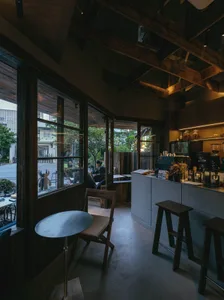 Interior view of a modern Taipei cafe featuring dark reclaimed wood ceiling beams, a concrete service counter, wooden stools, and large glass windows overlooking a tree-lined street.