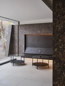 Modern minimalist lobby featuring raw OSB panel walls, dark stone feature wall with backlighting, and two round glass-top nesting coffee tables on light tiled floor.