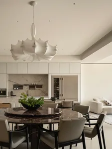 Modern minimalist dining area featuring a large round dark marble table, six beige upholstered chairs with black frames, and a striking white sculptural pendant lamp, with an open kitchen and living room in the background.