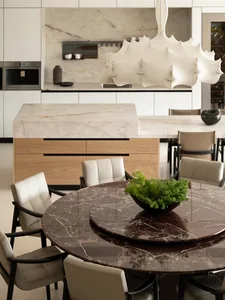 Modern dining area featuring a large round dark brown marble table surrounded by cream leather dining chairs, with a sculptural white pendant lamp hanging above and an open kitchen with light marble island in the background.