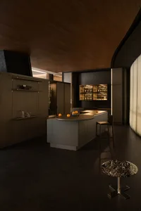 Modern dark-toned kitchen featuring a curved kitchen island, dark wood cabinetry, illuminated glassware display, and a black marble side table in the foreground.