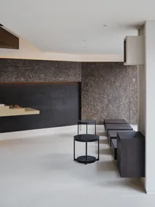 Modern minimalist living room featuring dark brown terrazzo accent wall and built-in bench, light wood floating console, black metal and terrazzo nesting tables, and white tiled floor.