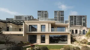 Front exterior view of a modern classical luxury villa constructed with light limestone, featuring tall columns, large glass windows, and a landscaped front garden with city high-rises in the background.
