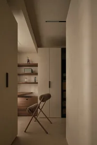 Minimalist dressing room featuring walnut floating shelves, built-in drawers, a tall beige wardrobe, and a wooden X-frame stool with bouclé upholstery.