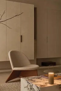 Modern minimalist living room featuring a beige upholstered lounge chair with wooden base, white marble coffee table, built-in beige cabinetry, and warm ambient lighting.