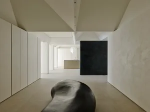 Long minimalist hallway with off-white micro-cement walls and floor, a large amorphous black sculptural bench in the foreground, and a dramatic dark textured accent wall at the end.