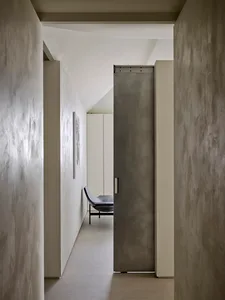 Narrow minimalist hallway with textured concrete walls, a large grey pivot door, and a black leather chaise lounge visible in the background under a sloped ceiling.