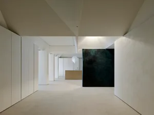 Modern minimalist interior hallway with white micro-cement walls and floor, leading to a kitchen island, featuring a large dark textured accent wall and a white pendant lamp.