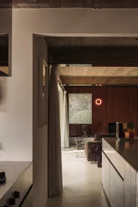 Interior view of a modern living room featuring a dark walnut wood accent wall, exposed concrete ceiling, brown bouclé sofa, and a glowing red circular wall light.