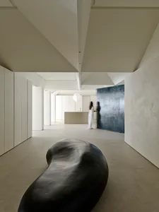 Minimalist interior hall featuring a large organic black sculptural bench in the foreground, textured micro-cement walls, high geometric ceiling, and a woman standing near a kitchen island in the background.