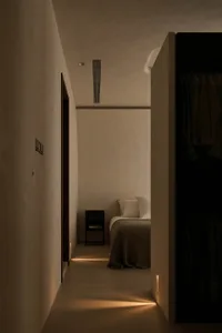 View down a narrow hallway into a minimalist bedroom featuring textured plaster walls, warm linear LED lighting at floor level, a low black nightstand, and a bed with textured brown throw blanket.