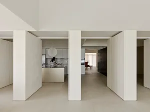 View from a hallway into a minimalist open-plan apartment featuring micro-cement walls, a central kitchen island, and a pendant light, with a woman cooking in the background.