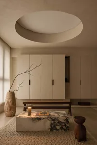 Minimalist living room featuring a large Calacatta marble coffee table, low wooden bench, sculptural dark wood stool, textured beige rug, built-in neutral cabinetry, and a dramatic circular recessed ceiling light.