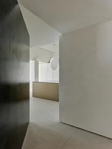 View from a dark hallway into a bright minimalist kitchen featuring micro-cement walls, concrete floor tiles, and a large white sculptural pendant lamp hanging over a metallic island.