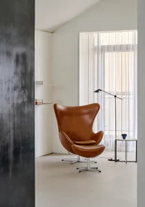 Brown leather Egg Chair with matching ottoman placed in a bright minimalist room with sheer curtains and a black floor lamp