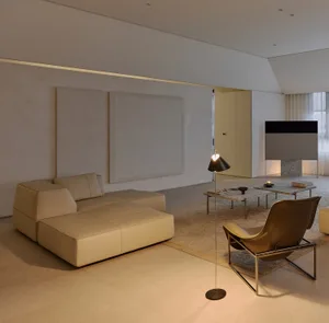 Minimalist living room featuring a large beige modular sofa, brown leather lounge chair, floor lamp, and expansive micro-cement walls with warm ambient lighting.