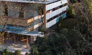 High-angle view of a modern multi-level house with corrugated metal cladding, wooden structural beams, and an open terrace with round wooden tables, surrounded by dense trees and foliage.