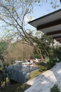 現代風戶外露台以耐候鋼結構與白色餐桌椅組成，俯瞰鬱鬱蔥蔥的森林山谷，有人正在樹下用餐。
