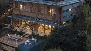 Night view of a modern two-story villa featuring extensive wooden decks, a lower-level Cor-Ten steel container-like structure with white outdoor furniture, and lush surrounding trees.
