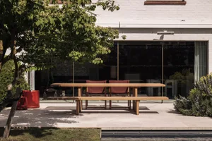 Modern outdoor dining area featuring a long solid teak wood table, matching bench, and two red upholstered chairs on a large-format stone patio in front of a white brick house with large glass doors.
