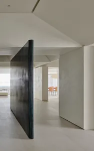 Modern minimalist interior featuring a tall patinated bronze pivot door leading to a bright open-plan living and dining area with neutral tones.