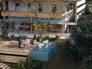 山間現代別墅鳥瞰圖，包含大型木製露台與用餐區、醒目的藍綠色耐候鋼平台，以及人們在樹木環繞的戶外空間休憩。