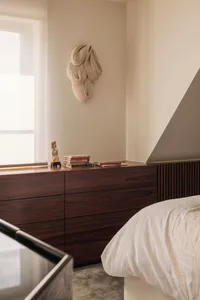 Modern minimalist bedroom corner featuring dark walnut built-in dresser, white bedding, a large textured beige fiber wall sculpture, and a small Chinese figurine on the windowsill.