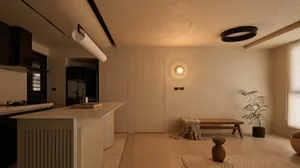 Modern minimalist open-plan kitchen and living room with warm beige microcement walls, light wood flooring, a central kitchen island with fluted detailing, wooden bench, potted tree, and circular ceiling lights.