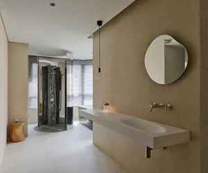 Modern minimalist bathroom featuring a long white floating vanity, irregular round mirror, textured micro-cement walls, and a dark marble walk-in shower in the background.