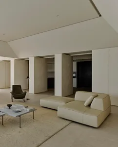Minimalist living room featuring a large beige leather modular sofa, low concrete coffee table on a textured rug, dark leather lounge chair, and high concrete ceilings with soft natural lighting.