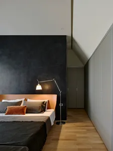 Modern minimalist bedroom with a large dark charcoal textured feature wall, tan leather upholstered headboard, layered grey and rust pillows, and a chrome articulated floor lamp.
