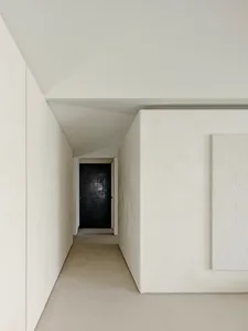Long narrow minimalist hallway with white walls, textured micro-cement finish, light concrete floor and a black door at the end