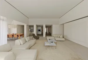Bright and airy open-plan living and dining area featuring a large curved white leather sofa, marble coffee tables, a sculptural dark green lounge chair, and a wooden dining table with terracotta chairs in the background.