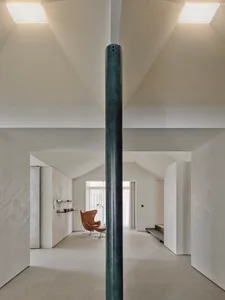 Modern minimalist living room with high sloped concrete ceiling, large aged green steel pillar in foreground, brown leather Egg chair in background, and soft natural light.