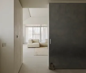View from a concrete hallway into a bright minimalist living room featuring off-white modular sofa, large windows with sheer curtains, and micro-cement walls.