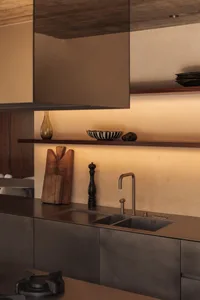 Modern minimalist kitchen featuring a brushed stainless steel countertop and double sink, dark cabinetry, floating walnut open shelf with decorative objects, and warm ambient lighting.