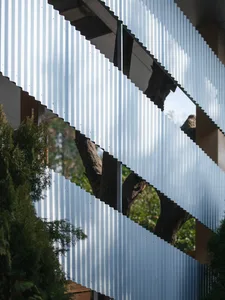 Close-up architectural detail of a modern building facade featuring angled corrugated metal panels that reflect green trees and sky.