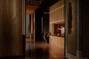 View into a luxurious private dining room featuring layered textured stone walls, dark marble accents, warm ambient lighting, and a chef preparing behind a stone counter.